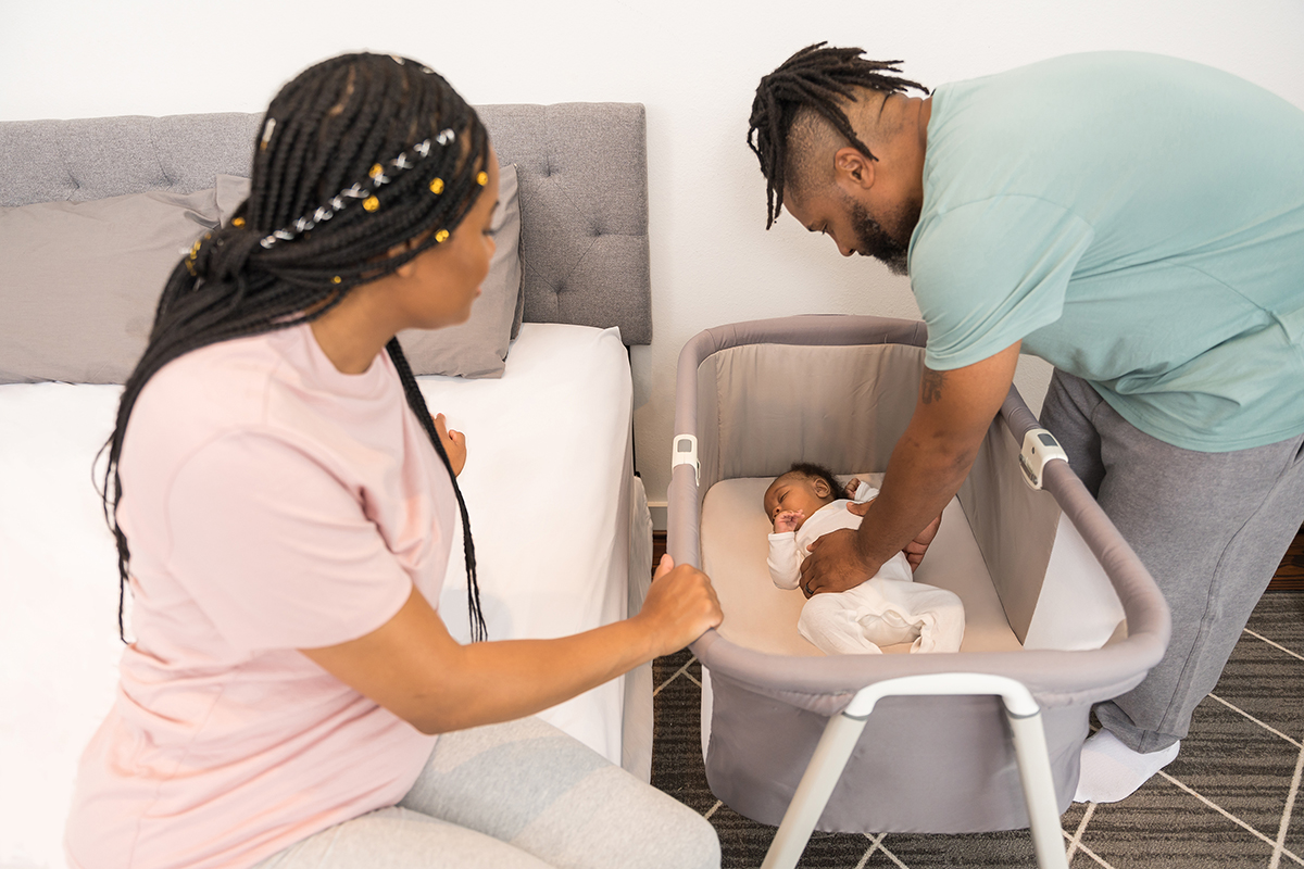 new parents putting their baby to sleep in a crib next to their bed