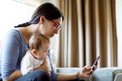 Texto alternativo: Una madre sentada sosteniendo a su bebé y mirando su celular y una computadora portátil.